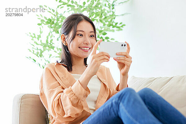 Asiatische Frau sitzt auf der Couch und macht ein Selfie mit ihrem Handy