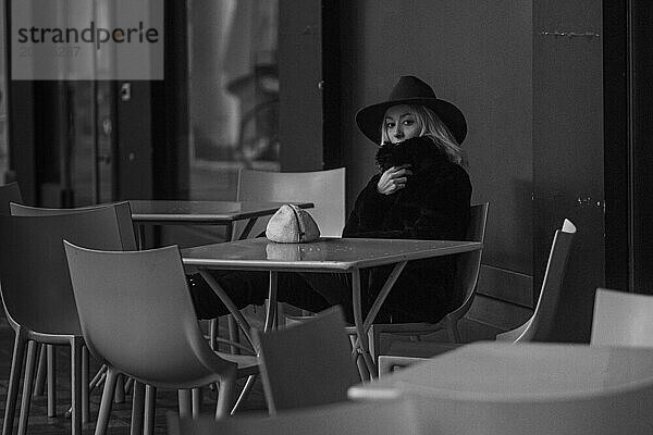 Schwarz Weiß Foto einer nachdenklichen Frau mit Hut  die an einem Tisch in einem Straßencafé sitzt