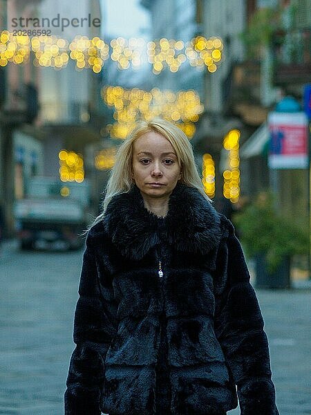 Fokussierte traurige blonde Frau im Pelzmantel  die abends auf einer Straße in der Stadt steht  mit Bokeh Lichtern