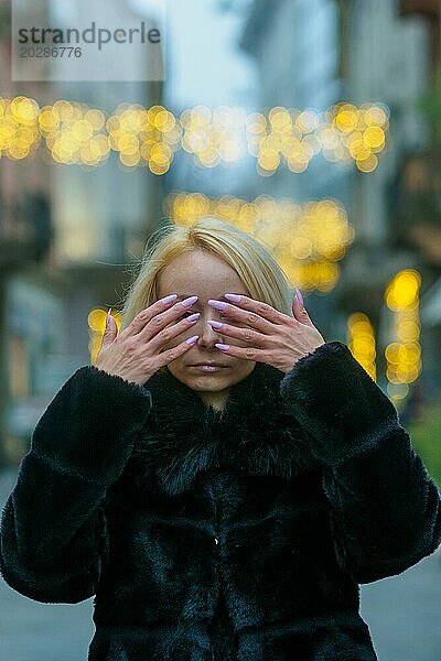 Eine blonde Frau in einem schwarzen Mantel bedeckt ihre Augen  mit abendlichen Bokeh Lichtern hinter ihr