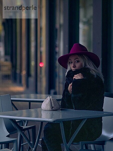 Frau mit magentafarbenem Hut und schwarzem Mantel  nachts im Freien sitzend  im Hintergrund die Lichter der Stadt