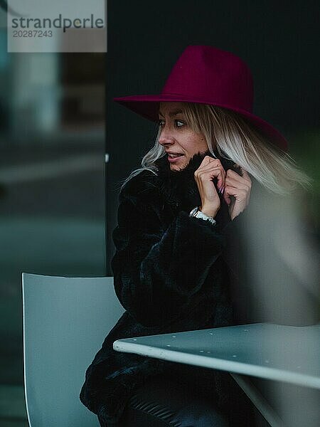Frau mit lilafarbenem Hut sitzt drinnen  lächelt und schaut in einer gemütlichen Umgebung weg