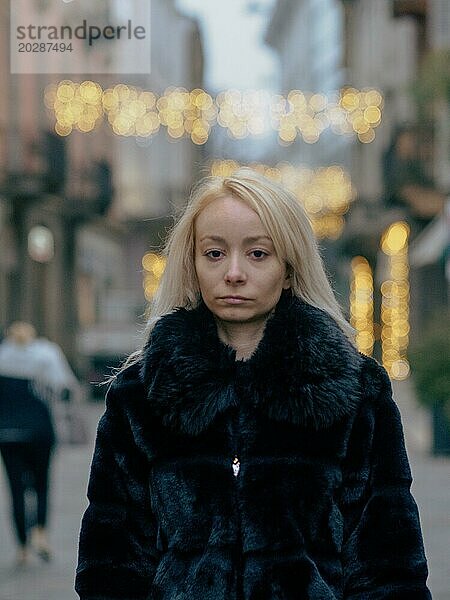 Eine nachdenkliche Frau in einem Pelzmantel steht auf einer Straße in der Stadt mit Abendlichtern mit Bokeh Effekt