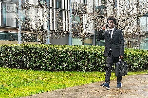 Afrikanischer junger Geschäftsmann beim Pendeln  während er in einem öffentlichen Stadtpark mit seinem Handy spricht