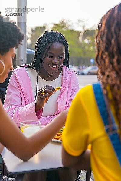 Vertikales Foto von drei jungen afrikanischen Freundinnen beim Essen von Nachos in einem Straßenrestaurant