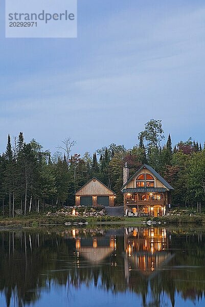Beleuchtete Doppelgarage und handgefertigtes zweistöckiges Blockhaus aus Fichtenholz mit Feldsteinschornstein und grünem Blechdach am Rande eines Sees in der Herbstdämmerung  Quebec  Kanada  Nordamerika