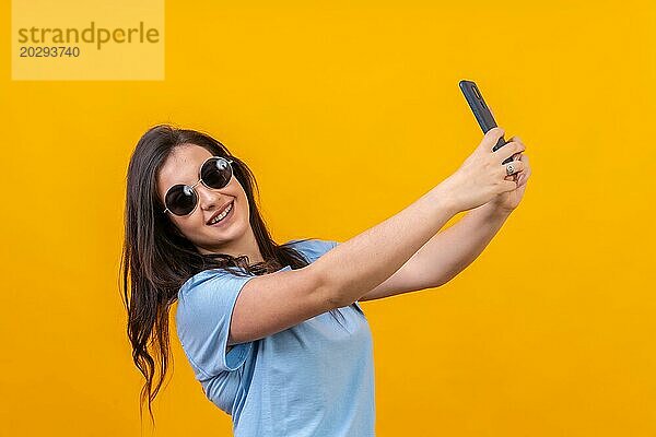 Studio Portrait mit gelbem Hintergrund einer schicken Frau mit Sonnenbrille  die ein Selfie macht und in die Kamera lächelt