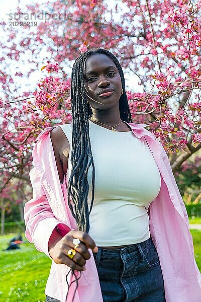 Vertikales Porträt einer jungen afrikanischen Frau in Freizeitkleidung  die neben einem blühenden rosa Baum posiert