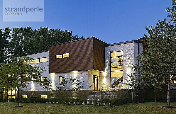 Beleuchteter beiger Stein mit brauner Zedernholzverkleidung modernes Haus im kubistischen Stil mit begrüntem Vorgarten in der Abenddämmerung im Sommer  Quebec  Kanada  Nordamerika