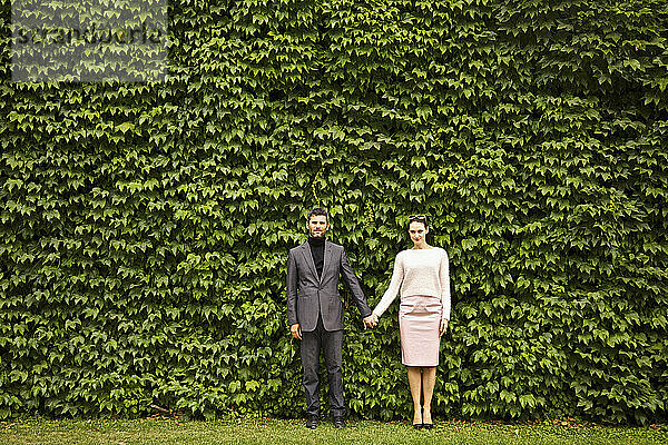 Paar hält sich an den Händen und steht vor einer grünen Hecke