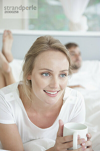 Frau liegt auf dem Bett und hält eine Tasse in der Hand