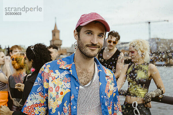 Porträt eines lächelnden schwulen Mannes im geblümten Hemd mit nicht-binären Freunden  die im Hintergrund Spaß haben