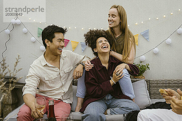 Fröhliche männliche und weibliche Freunde lachen zusammen  während sie auf dem Sofa auf einer Party auf dem dekorierten Balkon sitzen