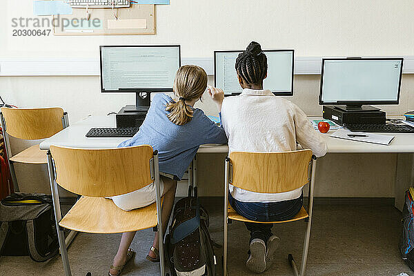 Rückansicht eines Mädchens  das einer Freundin beim Computergebrauch hilft  während es im Klassenzimmer der Schule auf einem Stuhl sitzt