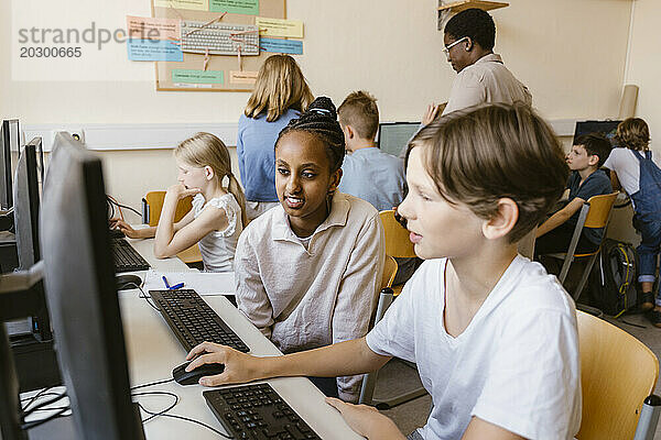 Lächelndes Mädchen sitzt mit einem männlichen Freund am Computer  während es in der Schule im Klassenzimmer sitzt