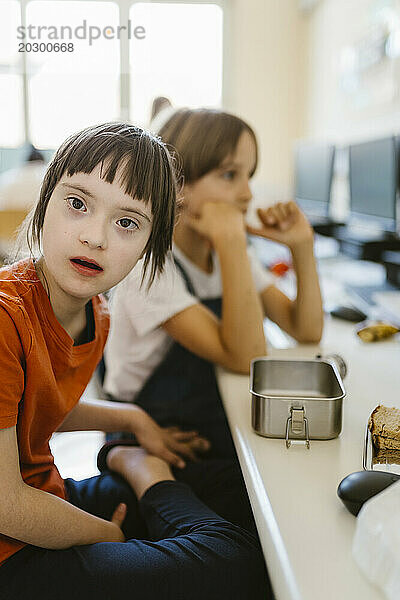 Porträt einer Studentin mit Down-Syndrom  die mit einer Lunchbox am Schreibtisch im Klassenzimmer sitzt