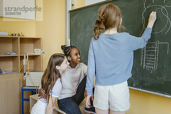 Mädchen haben Spaß mit einer Freundin  die im Klassenzimmer an die Tafel zeichnet