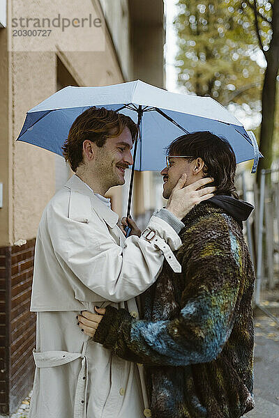 Seitenansicht eines jungen romantischen schwulen Paares  das unter einem Regenschirm auf der Straße steht