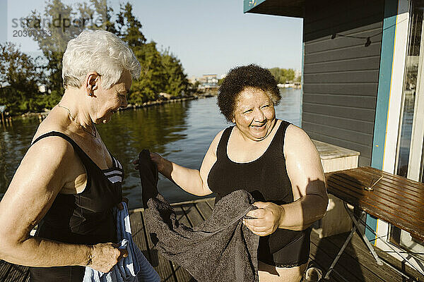 Fröhliche ältere Freundinnen halten ein Handtuch in der Hand  während sie an einem sonnigen Tag auf dem Hausboot stehen