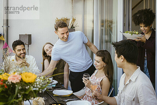 Lächelnde Männer und Frauen servieren Essen an Freunde  die während einer Party auf dem Balkon am Esstisch sitzen