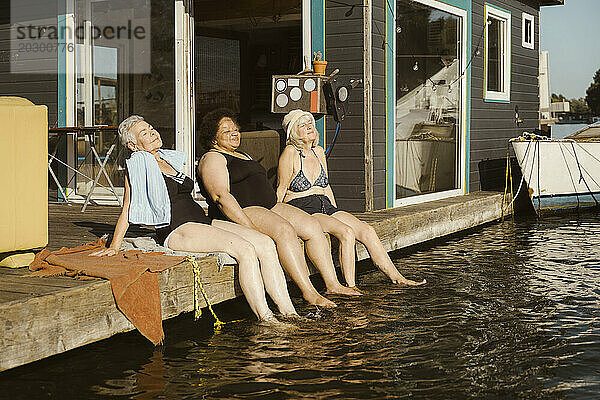 Lächelnde ältere Freundinnen tauchen ihre Beine ins Wasser  während sie sich im Urlaub auf dem Hausboot entspannen
