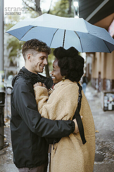 Seitenansicht eines glücklichen  gemischtrassigen Paares  das unter einem Regenschirm auf der Straße steht