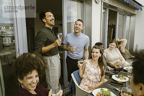 Eine Gruppe fröhlicher Freunde lacht  während sie auf dem Balkon eine Dinnerparty feiert