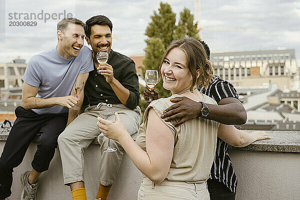 Porträt einer lächelnden Frau  die über die Schulter schaut  während sie mit männlichen Freunden auf der Dachterrasse Getränke genießt