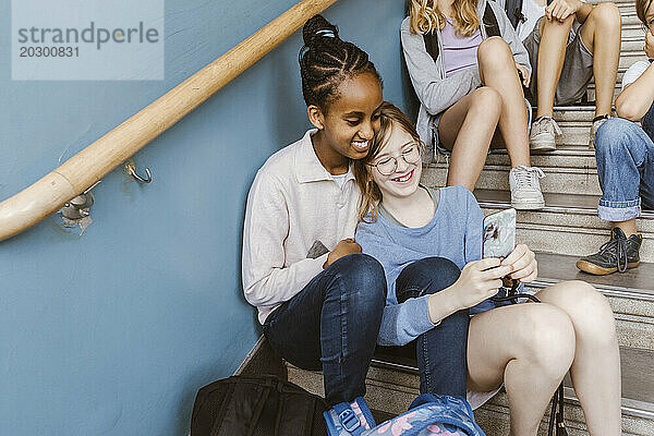 Fröhliche Freundinnen  die ihr Smartphone teilen  während sie in der Schule auf der Treppe sitzen