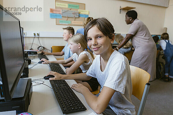 Porträt eines lächelnden männlichen Schülers  der mit Freunden am Schreibtisch im Computerklassenzimmer sitzt