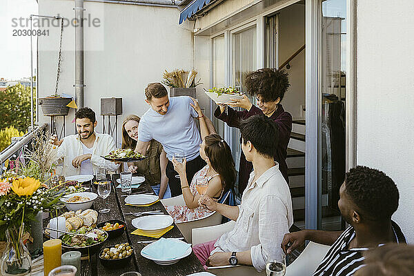 Glückliche Männer und Frauen servieren Essen für eine Gruppe von Freunden  die während der Dinnerparty auf dem Balkon am Esstisch sitzen