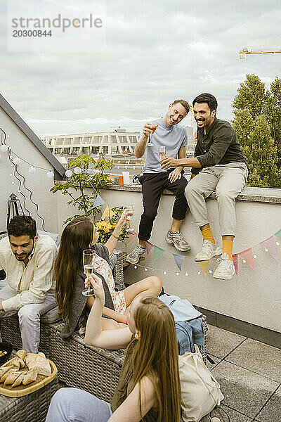 Lächelnde Männer stoßen mit Freunden auf Getränke an  während sie auf dem Balkon an der Wand sitzen