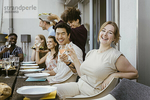 Porträt einer glücklichen jungen Frau  die ein Weinglas in der Hand hält  während sie mit einer Gruppe von Freunden bei einer Dinnerparty auf dem Balkon sitzt