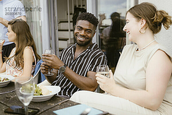Porträt einer lächelnden jungen Frau  die während der Dinnerparty auf dem Balkon mit einer Freundin am Esstisch etwas trinkt
