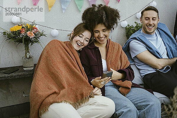 Fröhliche Freundinnen  die in eine Decke gehüllt sind und ihr Smartphone auf einer Party mit Freunden auf dem Balkon teilen