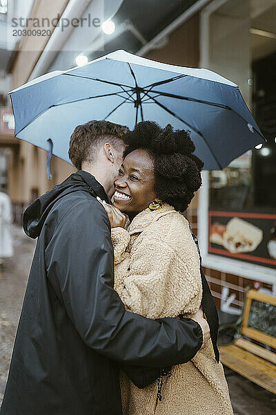 Freund umarmt glückliche gemischtrassige Freundin unter Regenschirm auf der Straße