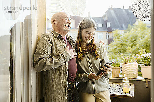 Glücklicher älterer Mann lacht  während er mit seiner Enkelin im Teenageralter steht  die ihr Smartphone auf dem Balkon hält