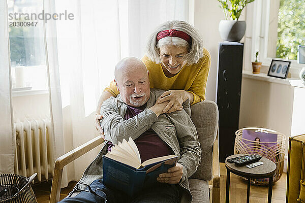 Glückliche ältere Frau mit dem Arm um den Mann  der ein Buch liest  während sie zu Hause auf einem Stuhl sitzt