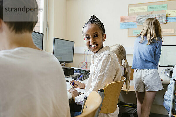 Seitenansichtporträt eines lächelnden Mädchens  das mit Freunden im Computerunterricht in der Schule auf einem Stuhl sitzt