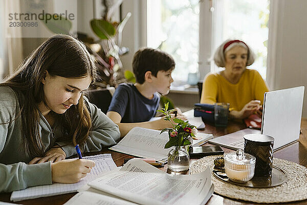Mädchen macht Hausaufgaben  während sie zu Hause mit Bruder und Großmutter sitzt