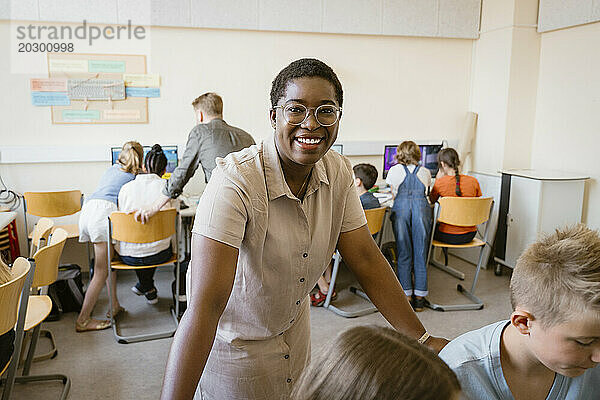 Porträt einer Lehrerin  die sich mit Schülern im Computerunterricht in der Schule auf Stühle lehnt