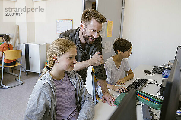 Lächelnder männlicher Lehrer  der einer Schülerin bei der Nutzung des Computers im Klassenzimmer der Schule hilft