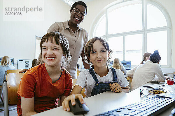Porträt eines lächelnden Mädchens neben einer Freundin mit Behinderung  die in der Schule im Computerunterricht sitzt