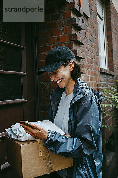 Fröhliche Zustellerin lacht  während sie mit dem Paket vor der Haustür steht