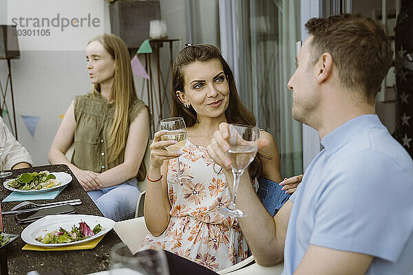 Lächelnde Frau hält Weinglas in der Hand und unterhält sich mit einem männlichen Freund während der Dinnerparty auf dem Balkon