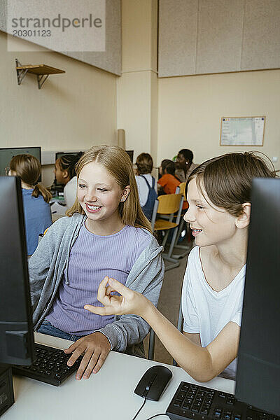 Lächelnder Junge  der mit einer Freundin am Computer spricht  während er im Klassenzimmer der Schule sitzt