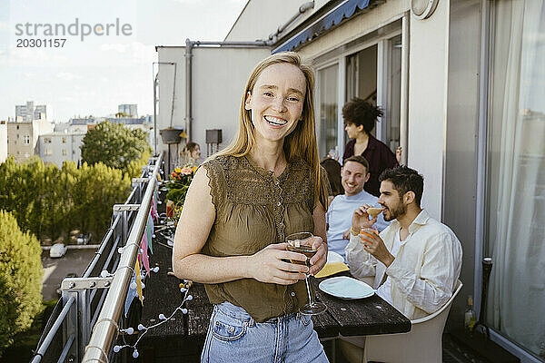 Porträt einer lächelnden Frau  die ein Glas Glas hält  während sie mit Freunden auf dem Balkon eine Dinnerparty feiert