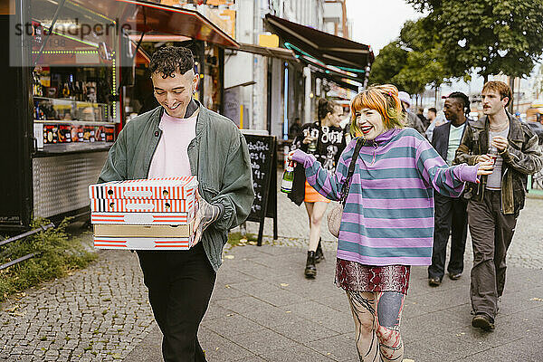 Lächelnde nicht-binäre Person hält einen Stapel Pizzakartons in der Hand  während sie mit Freunden auf der Straße in der Stadt spazieren geht