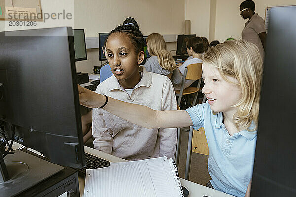 Mädchen zeigt auf Computermonitor  während sie mit einer Freundin im Klassenzimmer der Schule sitzt