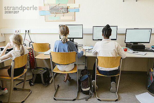 Rückansicht gemischtrassiger Studentinnen  die Computer benutzen  während sie im Klassenzimmer der Schule auf Stühlen sitzen